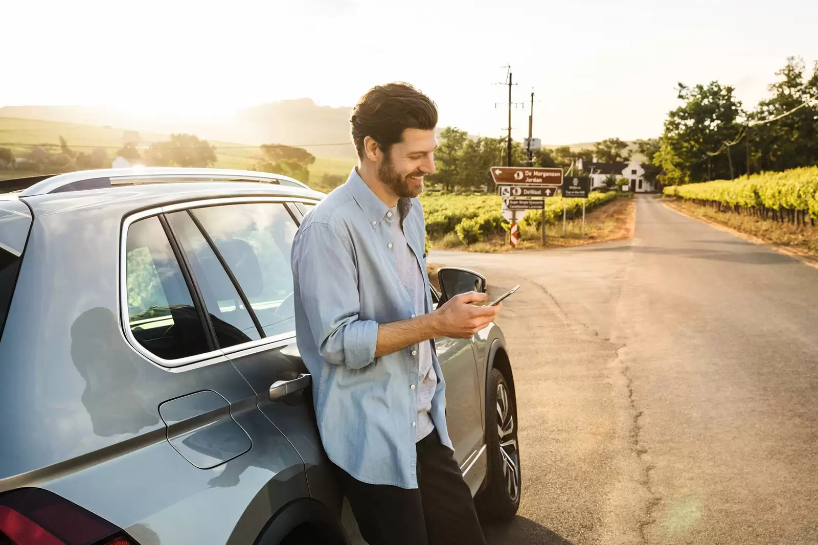 Hombre joven minrado en móvil apoyado en un coche aparcado en una carretera rural